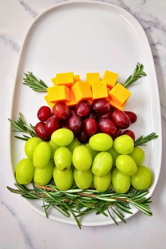 A Christmas fruit display in progress with rows of green and red grapes topped with bright cheddar cheese cubes and fresh rosemary sprigs, arranged in a tree shape on a white oval platter.