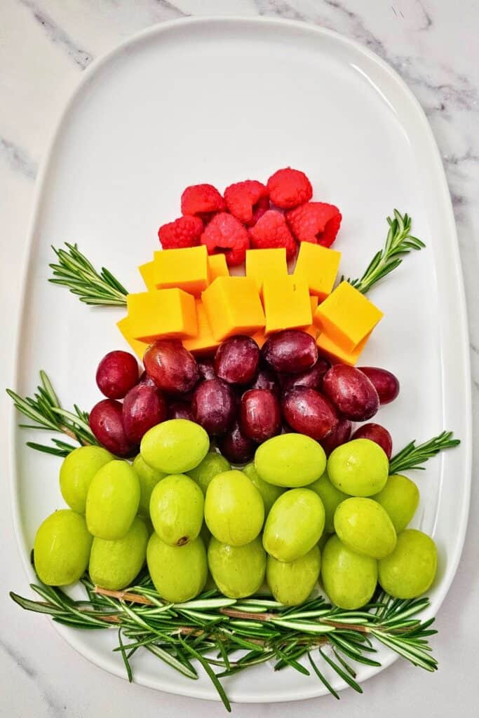 Overhead view of process shot of A fruit platter for Christmas arranged in a tree shape on a white oval tray, with layers of green grapes, red grapes, cheddar cheese cubes, and fresh raspberries accented by rosemary sprigs.