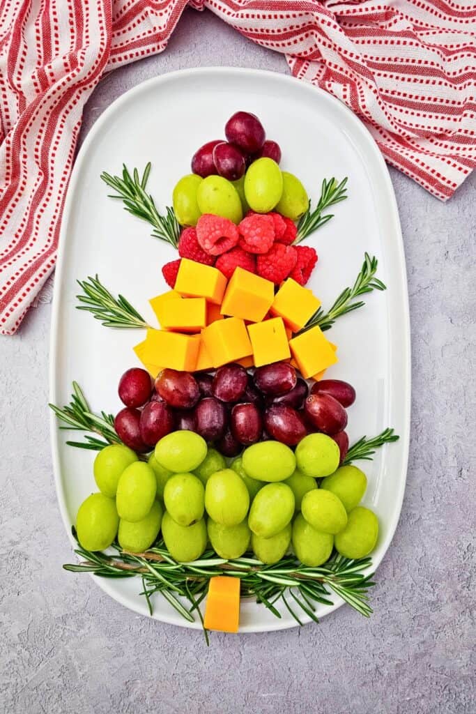 A festive Christmas tree made with layers of green and red grapes, raspberries, cheddar cheese cubes, and fresh rosemary on a white oval platter is a fun way to serve fruit tray ideas for Christmas gatherings.