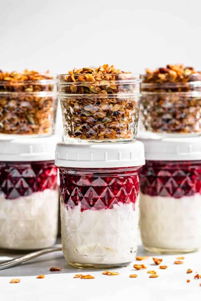 Three small mason jars filled will yogurt & berry sauce, with mini mason jars of granola.