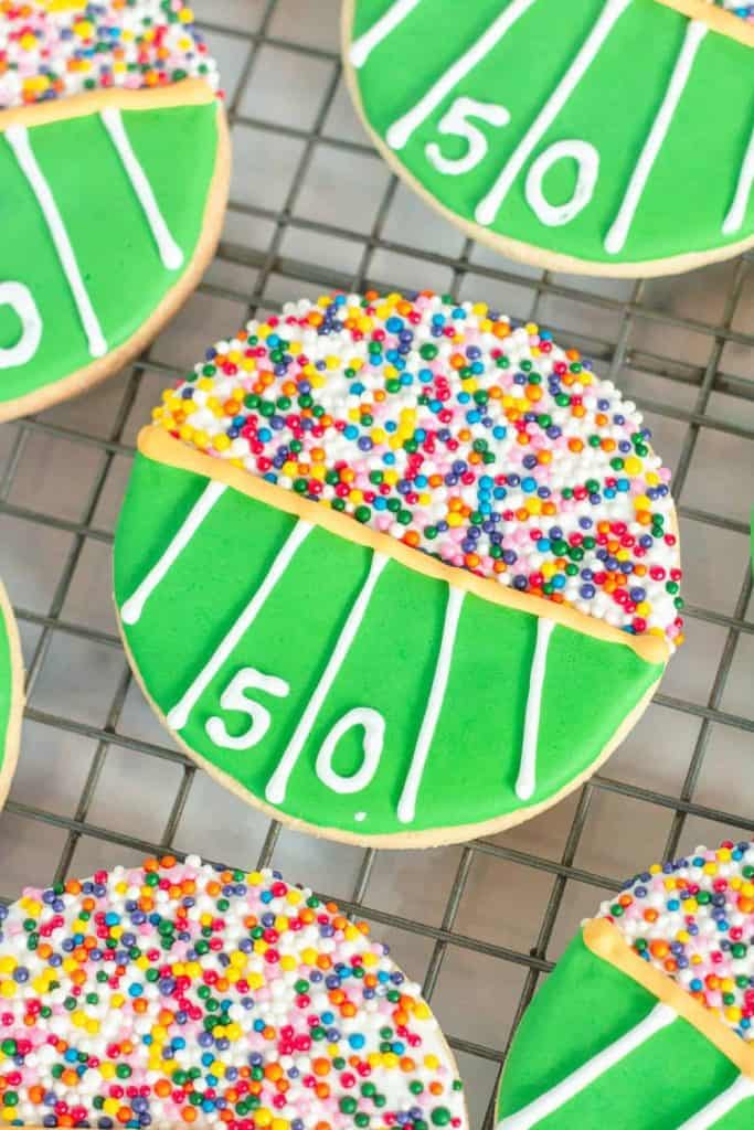 Round sugar cookies decorated to resemble football stadiums, with green icing fields marked with white yard lines and the number “50” at midfield. The top half of each cookie is covered in rainbow sprinkles, mimicking a crowd of colorful fans in the stands.