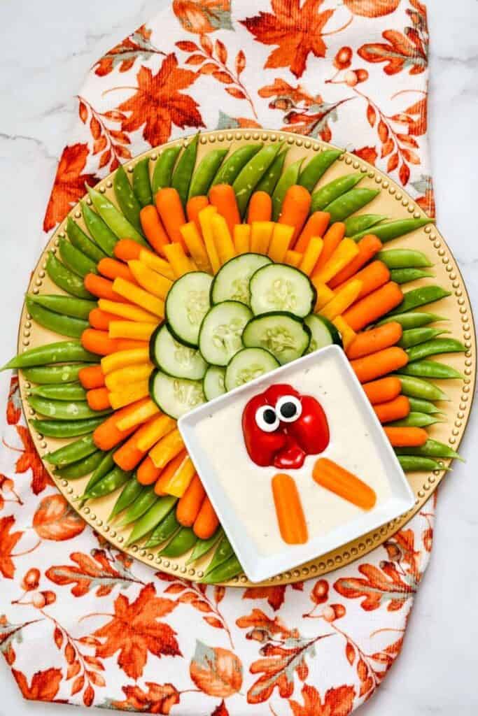 Overhead virw of a turkey vegetable tray on a round plate with green snap peas, baby carrots, yellow bell pepper strips, and cucumber slices arranged in colorful rows to resemble tail feathers. A bowl of creamy dip forms the body, with red bell pepper, edible eyes, and carrot pieces creating the turkey's face and wattle.