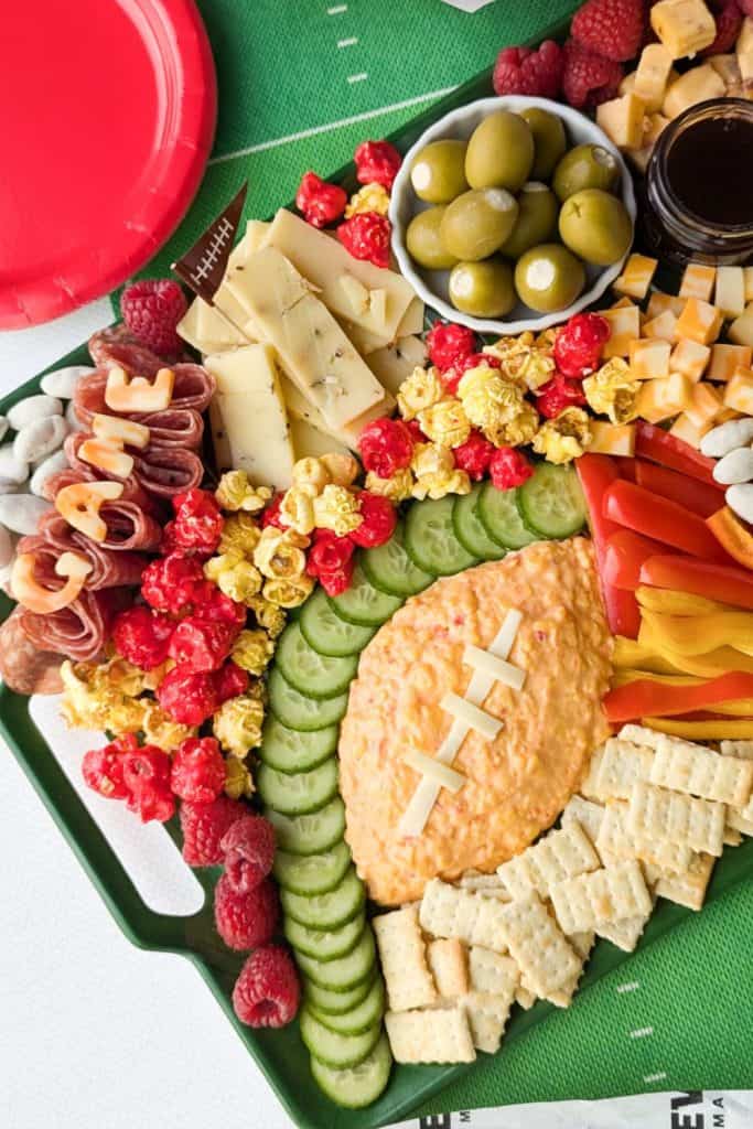 Game day charcuterie board arranged on a green football field tray with pimento cheese shaped like a football, topped with white cheese laces. Surrounded by sliced cucumbers, red and yellow bell peppers, popcorn, salami, cubed cheese, crackers, green olives, and raspberries.
