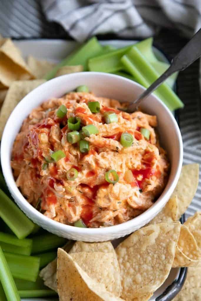 Crockpot buffalo chicken dip served in a white bowl, topped with hot sauce and chopped green onions. Surrounded by celery sticks and tortilla chips, this creamy and spicy dip is a game day favorite.