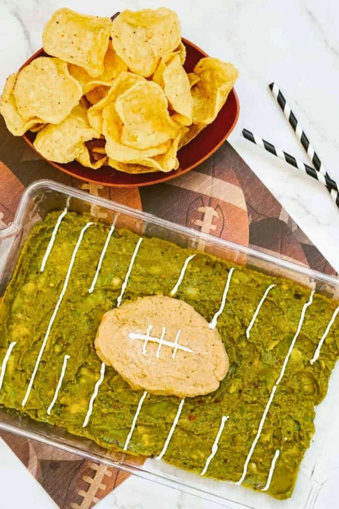 Game Day Guacamole Dip arranged in a glass dish to look like a football field, with sour cream lines as yard markers and a football-shaped refried bean topping in the center. Served alongside a bowl of tortilla chips, making it a festive tailgate snack.