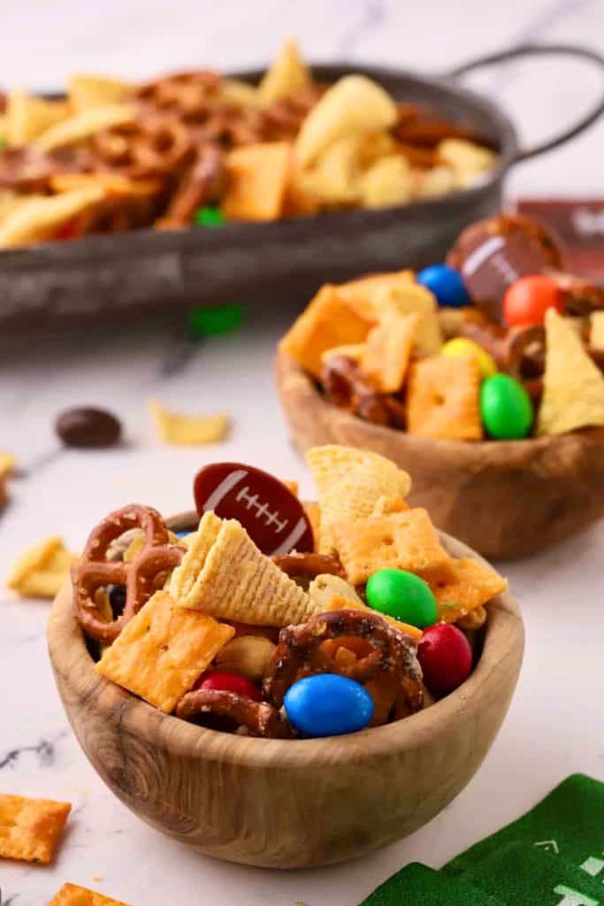 Game Day sweet and salty snack mix served in small wooden bowls, featuring pretzels, Chex mix, Cheez-Its, candy-coated chocolates, and Bugles. Mini football decorations are tucked into the mix, adding a festive touch.