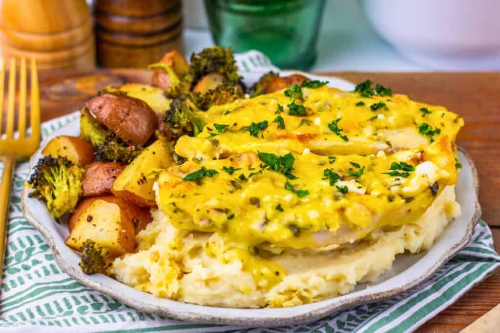 Close-up of garlic parmesan chicken served over mashed potatoes, topped with a golden cheese sauce and fresh parsley, alongside roasted baby potatoes and broccoli. The plate rests on a green and white striped napkin with a rustic wooden table beneath.