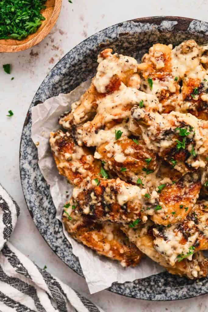 A bowl of crispy chicken wings generously coated in creamy garlic parmesan sauce, garnished with chopped parsley, served on parchment paper in a rustic stoneware dish.