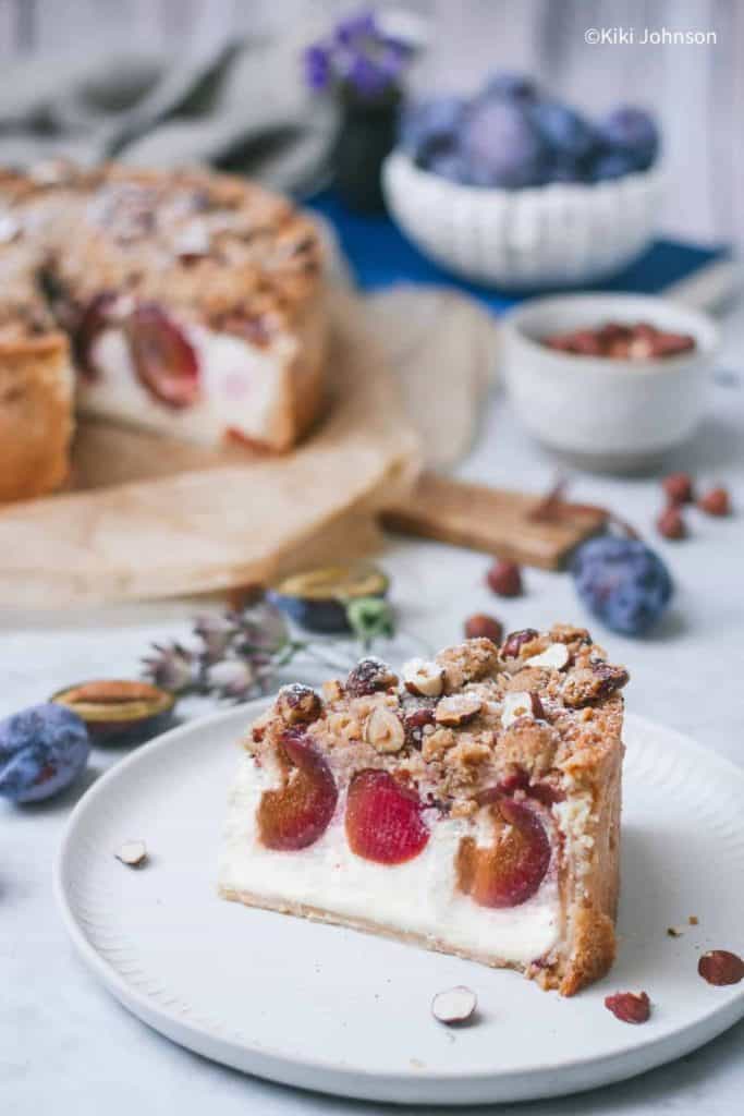 A slice of creamy plum cheesecake topped with chopped hazelnuts and a crumbly streusel layer, served on a white plate. Whole and halved plums, nuts, and a full cheesecake are in the background, creating a cozy, rustic fall dessert setting.