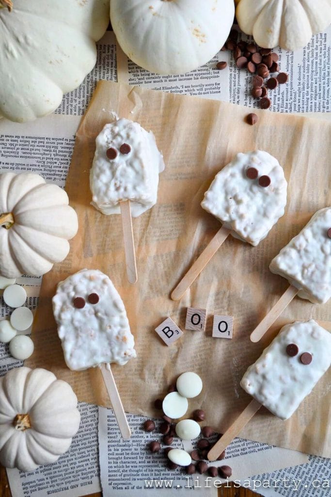 White chocolate-covered ghost-shaped Rice Krispie treats on wooden sticks are arranged on vintage book pages and parchment, decorated with chocolate chip eyes. Surrounded by white pumpkins, chocolate chips, candy melts, and Scrabble tiles spelling “BOO."