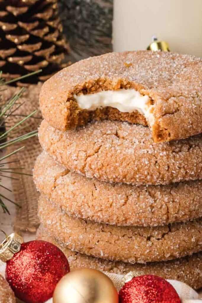 Stack of sparkly gingerbread cheesecake Christmas cookies, coated in sugar with a creamy cheesecake filling peeking from a bitten cookie on top, surrounded by pine branches and ornaments