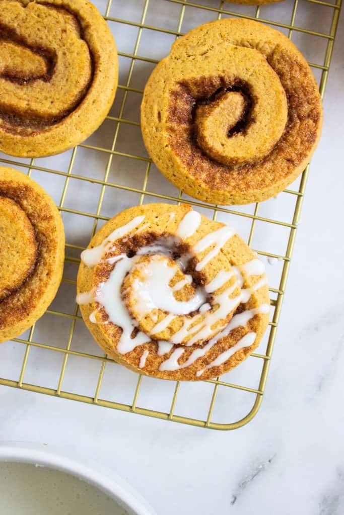 Pumpkin cinnamon roll cookies cooling on a wire rack, with one cookie freshly drizzled in glossy white glaze. The swirled centers and golden-brown edges highlight the soft, spiced texture of these fall treats.