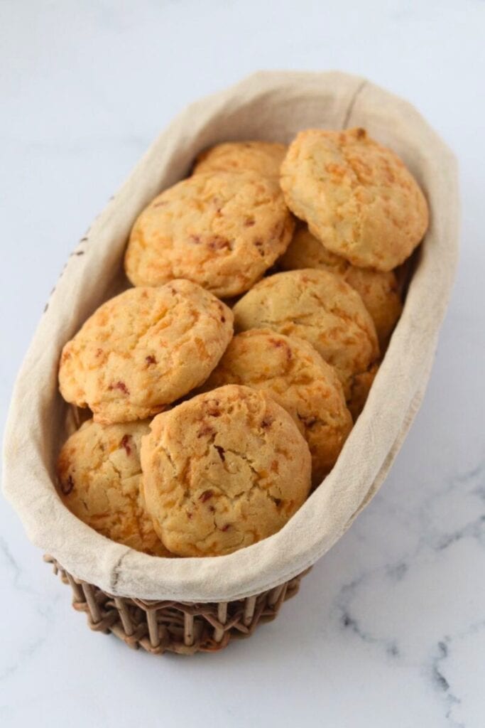 Basket of gluten free cheese and bacon biscuits with golden tops and a slightly cracked, tender texture. The biscuits are nestled in a fabric lined wicker basket, showing bits of melted cheese and small pieces of bacon throughout the perfect camping breakfast. 