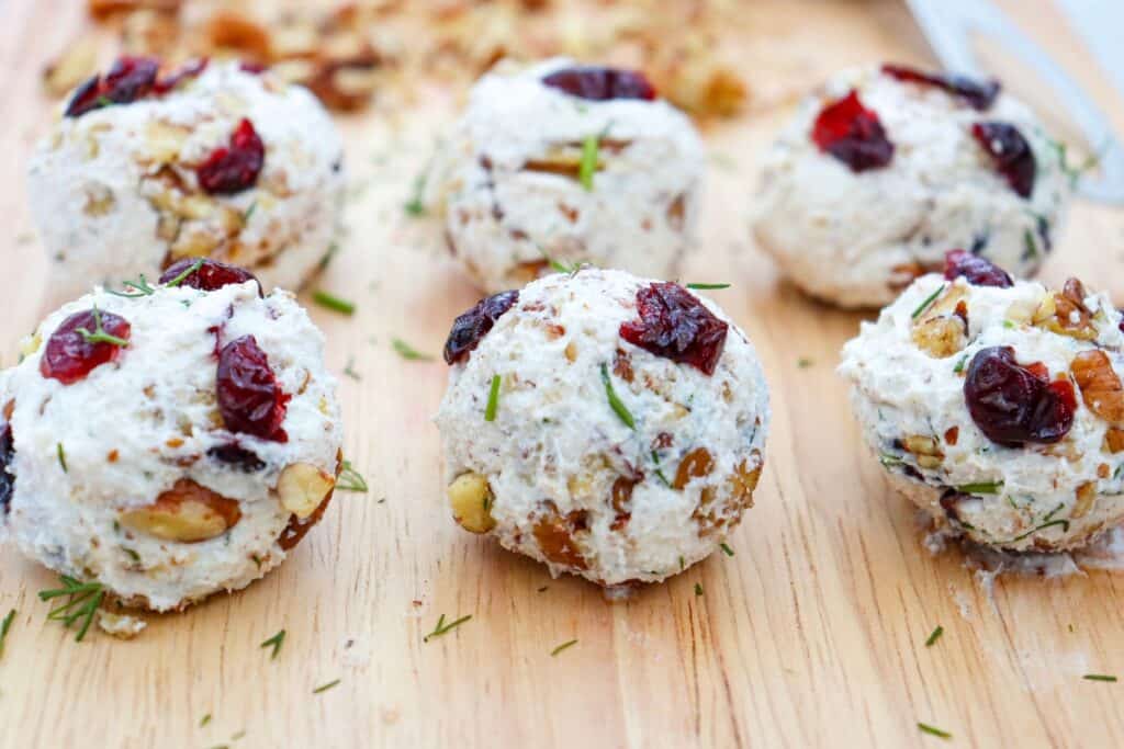 Six round goat cheese balls studded with dark red dried cranberries. Chopped pecans and flecks of fresh green dill cover the creamy white appetizers arranged on a wooden serving board.