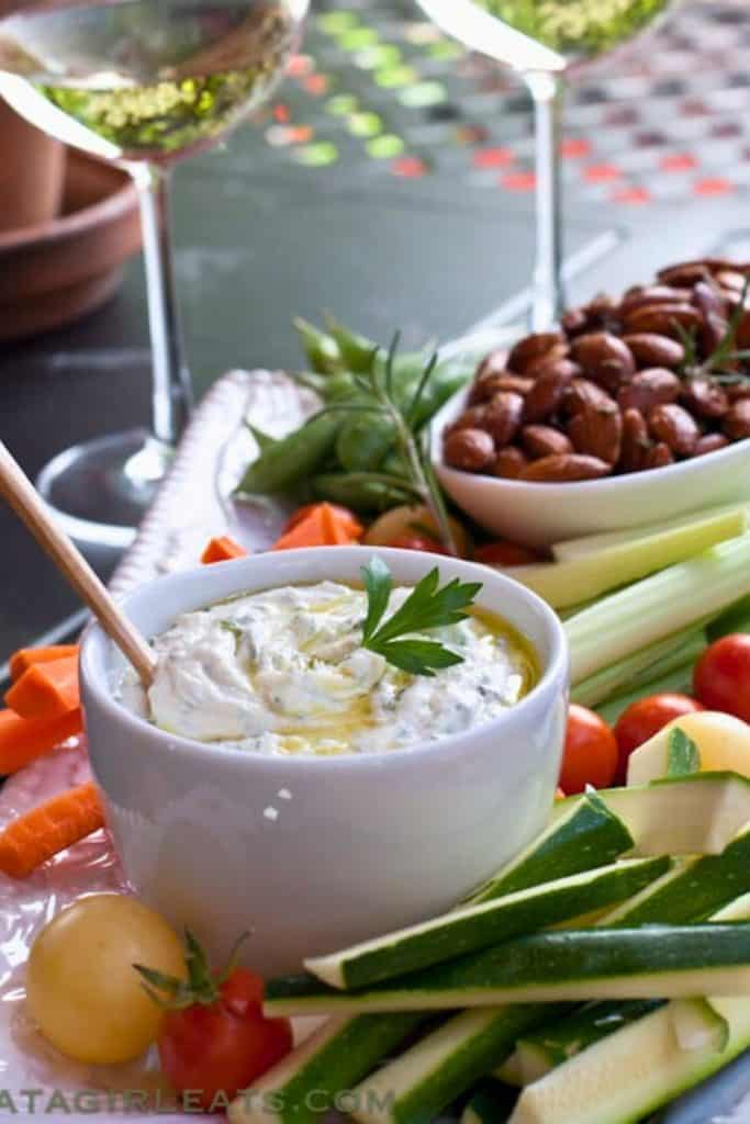 Creamy goat cheese dip in a white bowl, drizzled with olive oil and garnished with a fresh parsley leaf. Surrounded by an assortment of fresh vegetables, nuts, and wine glasses, making it an elegant appetizer spread.