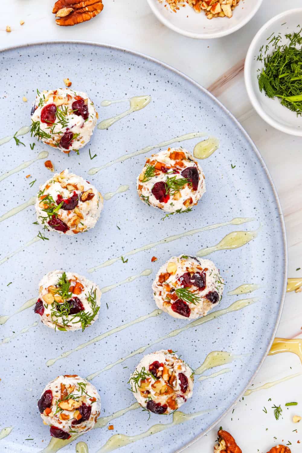 Over head view of seven round goat cheese truffles spaced across a large pale blue platter and coated with pieces of dark red dried cranberries, chopped brown pecans, and green dill. Thin lines of honey are drizzled across the platter, with small bowls of chopped pecans and fresh dill and a few whole pecans visible around the edges.
