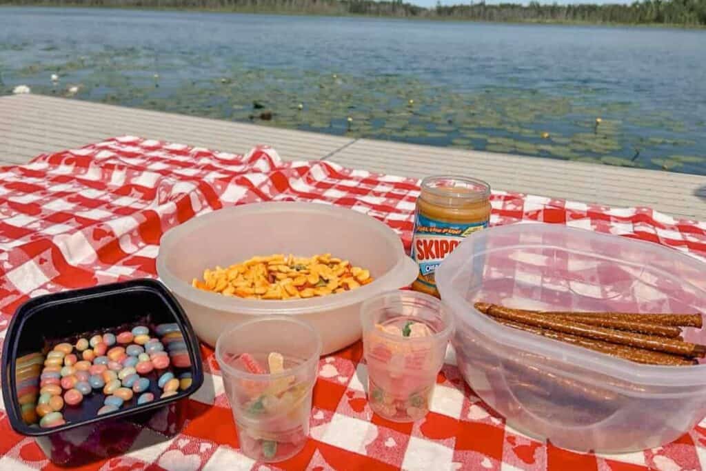 Picnic table with containers of peanut butter, pretzels, candy by a lake.