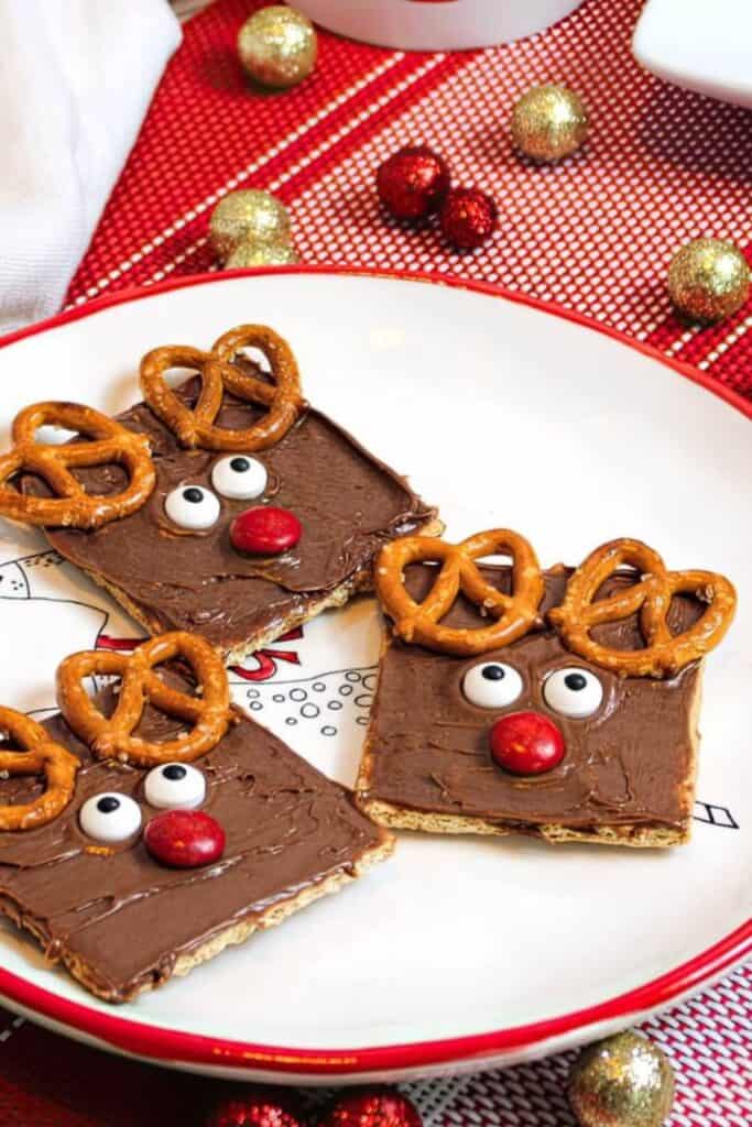 Square graham crackers are spread with chocolate frosting and decorated to look like reindeer using candy eyes, red M&M noses, and mini pretzel twists as antlers. These fun and festive no-bake treats are arranged on a holiday-themed plate surrounded by glittery ornaments.