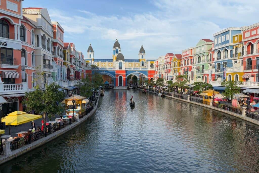 A colorful canal scene in Grand World Phú Quốc, Vietnam, shows pastel-hued European-style buildings lining both sides of a waterway. Gondolas glide through the canal, and pedestrians stroll beside café umbrellas and market stalls under a clear blue sky. 