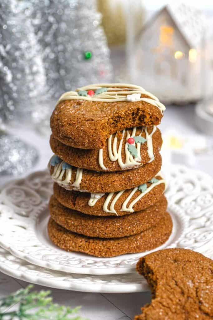 Thick ginger molasses cookies stacked on a white plate are decorated with white chocolate drizzle and tiny holly leaf sprinkles. One cookie has a bite taken out, showing its soft, chewy texture.