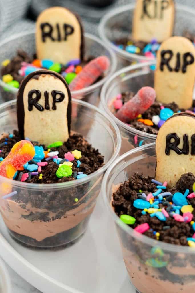 Clear plastic cups filled with layered chocolate pudding and crushed Oreo cookies, decorated with candy bones, sprinkles, gummy worms, and a cookie tombstone labeled "RIP" in black icing. These Halloween dirt cups resemble spooky graveyard scenes