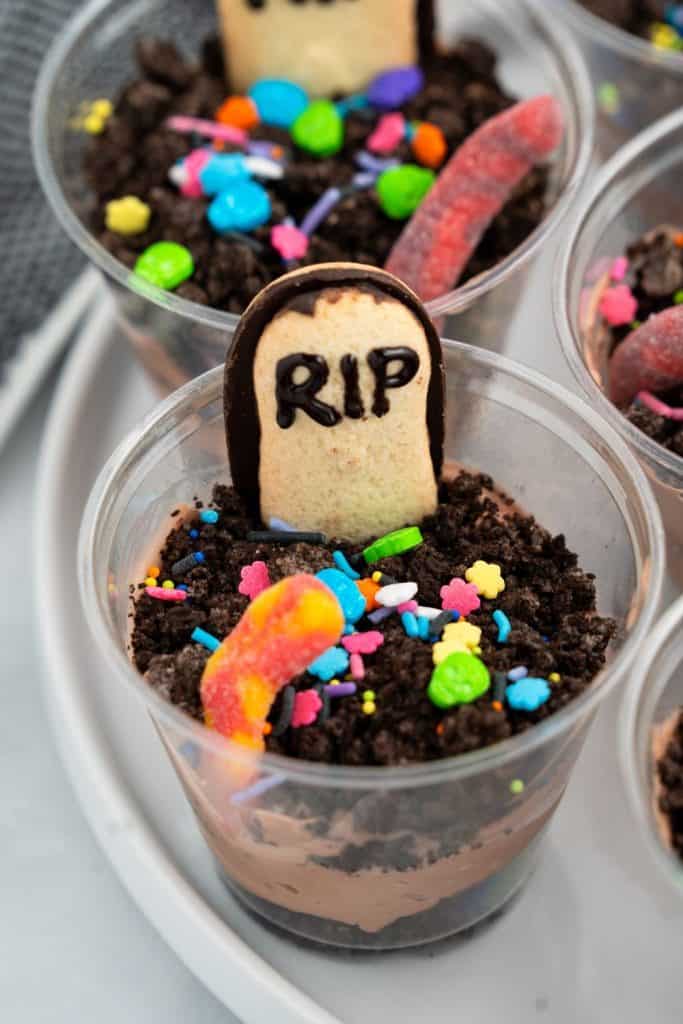 Plastic cup filled with layers of chocolate pudding and crushed chocolate cookies to resemble dirt, topped with a gummy worm, colorful Halloween-themed sprinkles, and a cookie shaped like a tombstone with “RIP” written in icing. The treat is styled to look like a mini graveyard, perfect for Halloween.