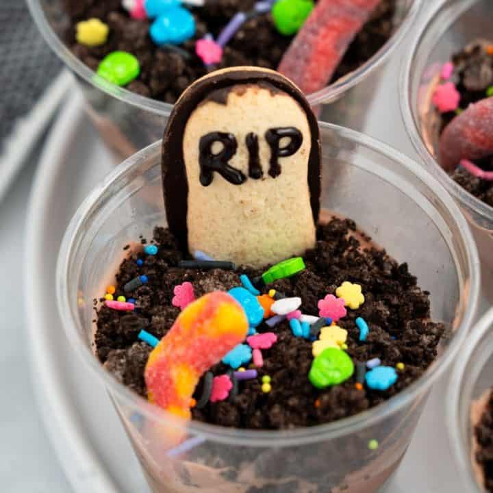 Plastic cup filled with layers of chocolate pudding and crushed chocolate cookies to resemble dirt, topped with a gummy worm, colorful Halloween-themed sprinkles, and a cookie shaped like a tombstone with “RIP” written in icing. The treat is styled to look like a mini graveyard, perfect for Halloween.