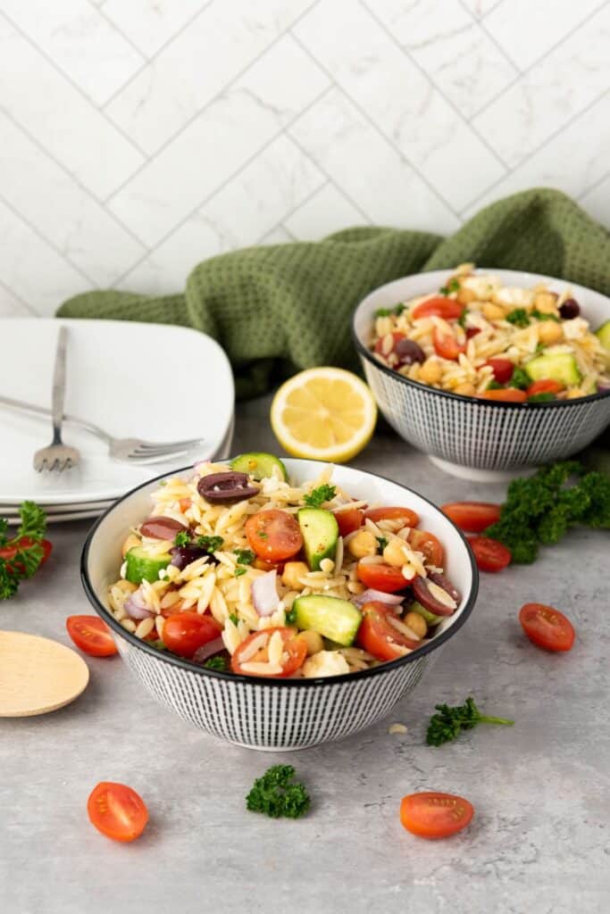 Two ceramic decorative bowls filled with Greek Orzo Pasta Salad beside a two plates with crossed forks on top.