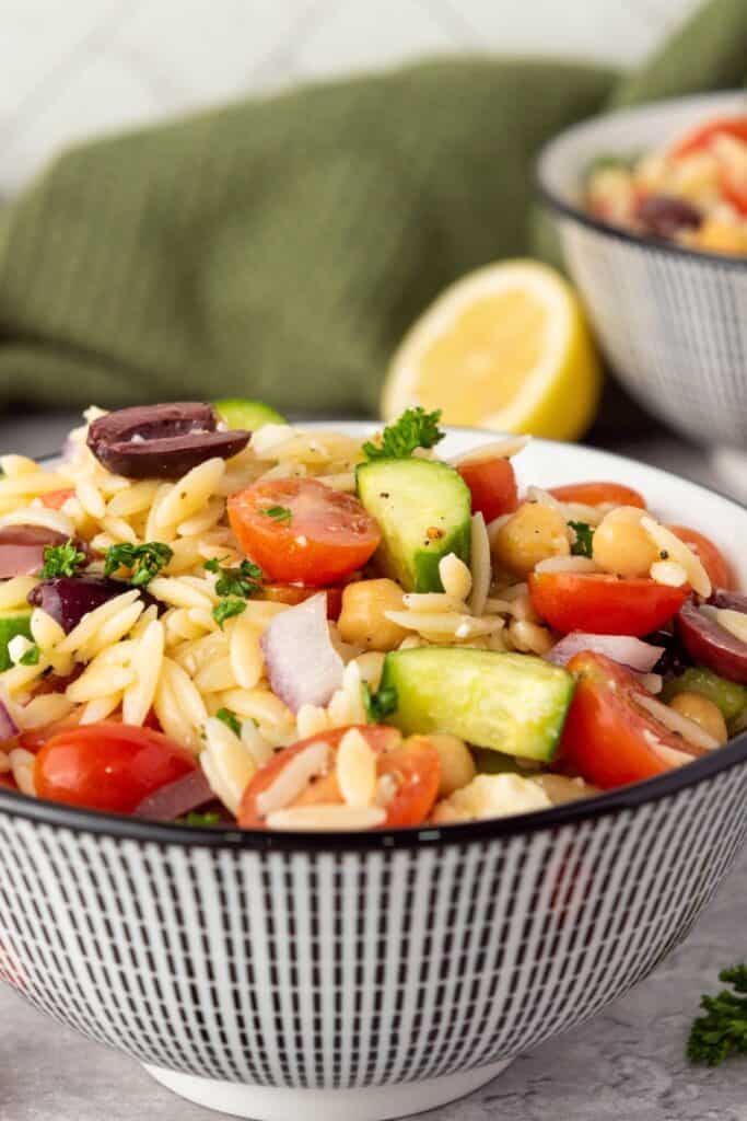Bowl of Greek orzo salad with orzo pasta chickpeas cherry tomatoes cucumber red onion kalamata olives and parsley tossed in a light lemon dressing. A lemon half and another bowl of the Mediterranean pasta salad sit in the background.