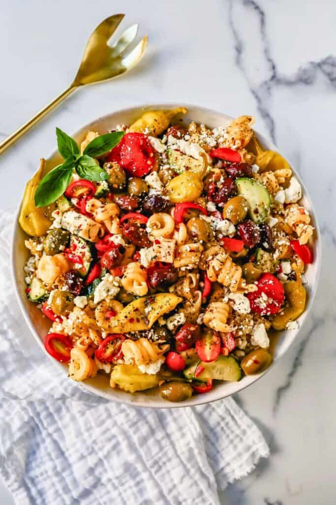 Vibrant Greek pasta salad in a white bowl, packed with rotini pasta, sliced cucumbers, cherry tomatoes, red peppers, kalamata and green olives, pepperoncini, and crumbled feta cheese. The salad is garnished with fresh basil and a sprinkle of herbs, with a gold serving fork and white cloth napkin beside it.