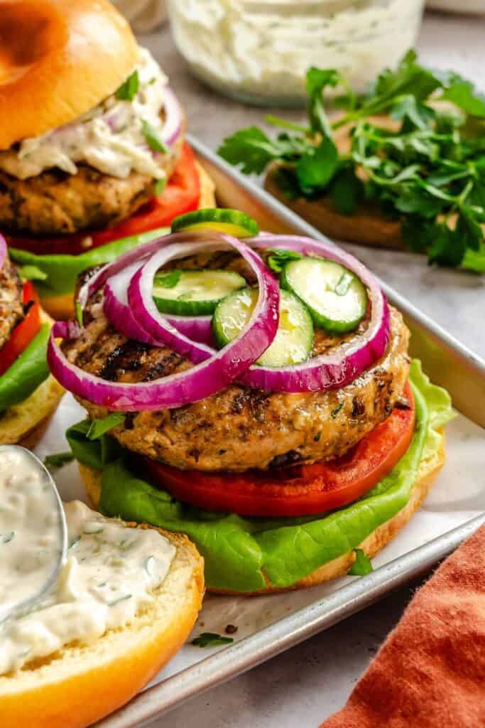Greek turkey burger served on a toasted bun with lettuce, tomato, grilled turkey patty, sliced cucumbers, and red onion. A creamy herb sauce is spread on the bun and extra burgers and fresh parsley are visible in the background, highlighting a fresh and flavorful Greek-inspired meal.