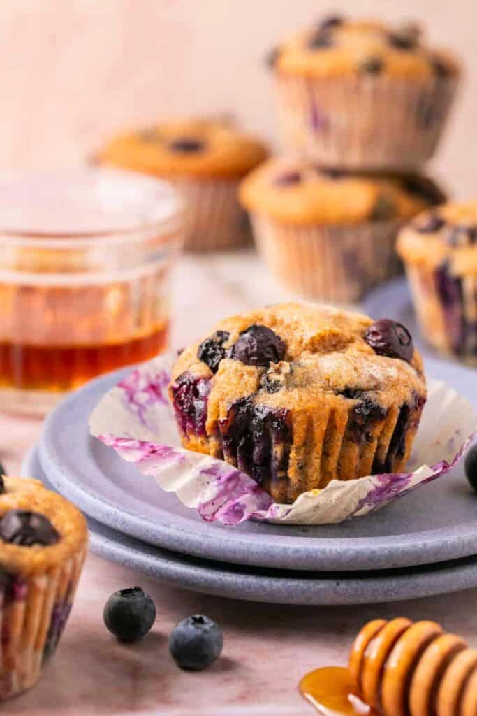 A Greek yogurt blueberry muffin sits unwrapped on a lavender plate, bursting with juicy whole blueberries and a golden, tender crumb. Fresh blueberries and a honey dipper with drizzled honey accent the scene, with more muffins and a glass of honey in the softly blurred background.