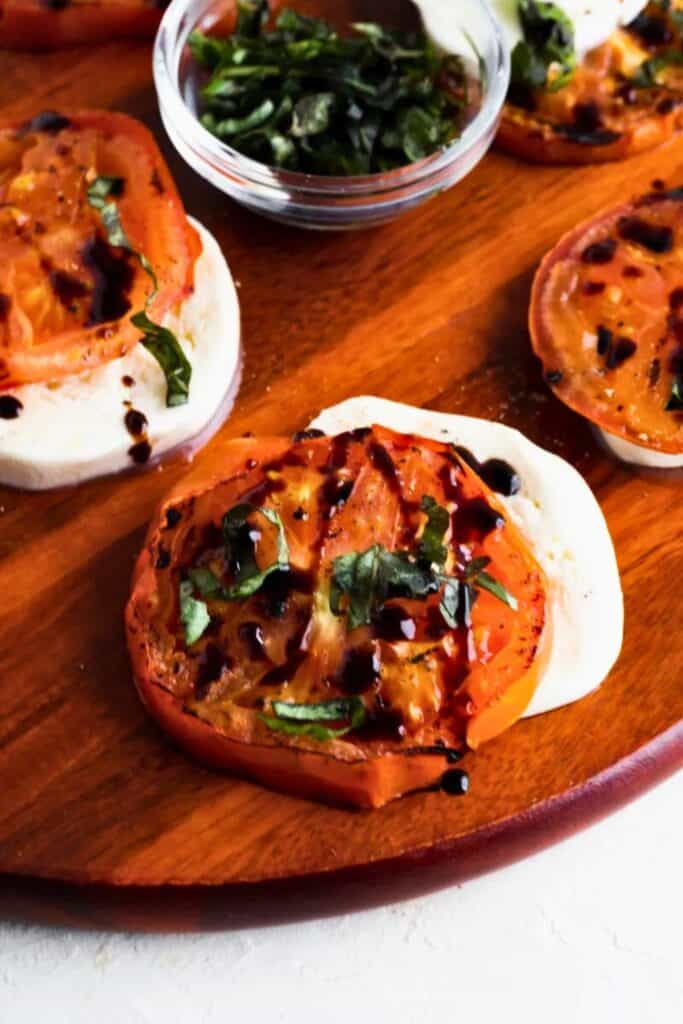 Grilled tomato slices layered over fresh mozzarella rounds, topped with chopped basil and drizzled with balsamic glaze, all arranged on a wooden serving board. A small glass bowl of extra basil garnish sits nearby.