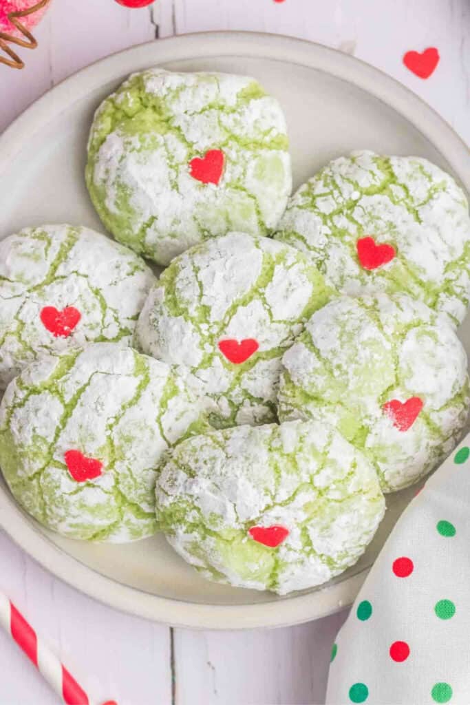 Green crinkle cookies dusted in powdered sugar are decorated with small red candy hearts in the center, resembling the Grinch’s heart.
