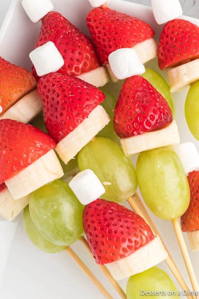 Grinch fruit kabobs made with green grapes, banana slices, strawberries, and mini marshmallows stacked on wooden skewers to resemble the Grinch wearing a Santa hat, perfect for a healthy holiday movie snack.