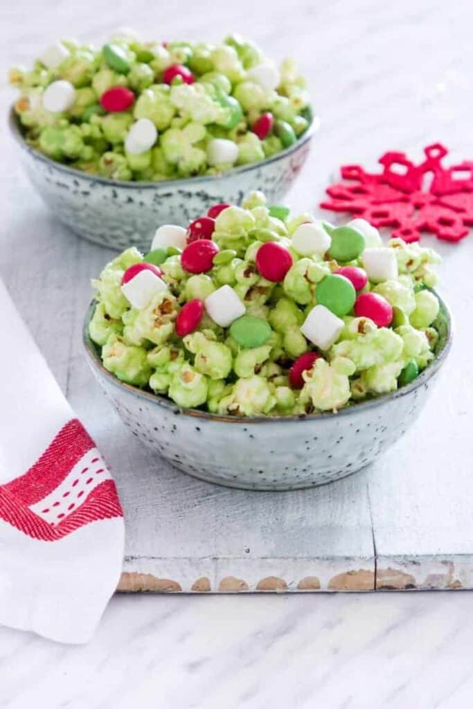 Two bowls of Grinch popcorn coated in green candy, mixed with red and green M&Ms and mini marshmallows, creating a colorful and festive sweet movie snack.