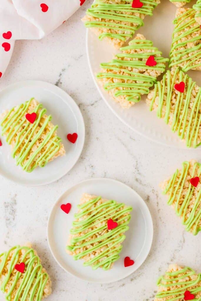 Grinch-themed Rice Krispie treats rectangles  drizzled with green icing and topped with one red candy heart each, served on white plates for a fun and festive addition to Christmas movie night snacks.