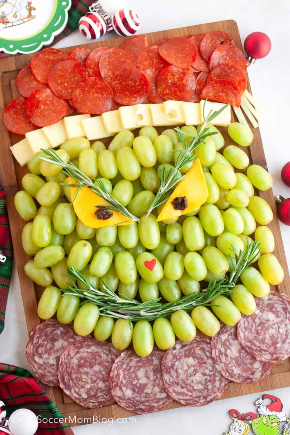 Grinch charcuterie board arranged with green grapes to form the Grinch face and pepperoni and salami to create his red Santa hat and collar. Cheese forms the hat trim and yellow eyes while rosemary sprigs create the eyebrows and curved smile.