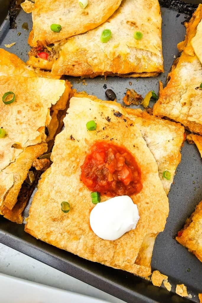 Golden baked quesadilla square in a sheet pan, topped with a dollop of sour cream, salsa, and a sprinkle of green onions. Its crispy exterior and simple, savory filling make it a kid-friendly quesadilla recipe perfect for easy family dinners.