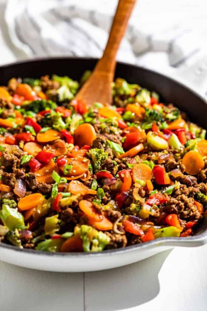 Quick ground beef dinner idea cooked in a skillet with browned ground beef, broccoli, carrots, red bell pepper, and onions tossed together with a wooden spoon. The colorful beef and vegetable mixture fills the pan for an easy one pan meal.