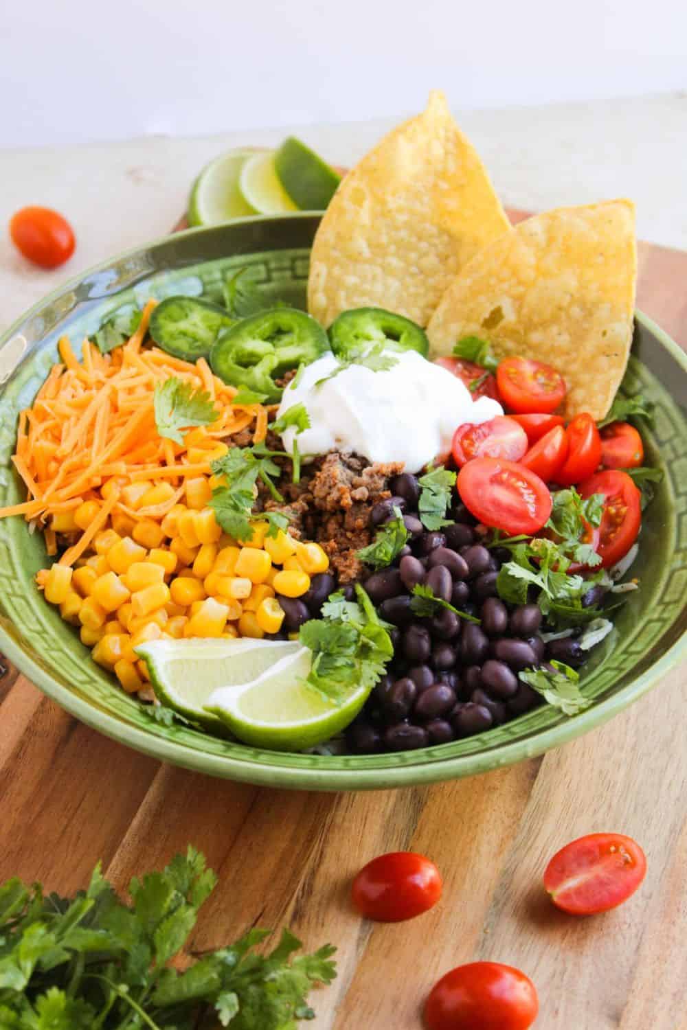 Colorful taco bowl topped with seasoned ground meat, black beans, corn, shredded cheddar cheese, sliced jalape&ntilde;os, cherry tomatoes, sour cream, cilantro, lime wedges, and tortilla chips. This quick and easy 30 minute summer dinner is served in a green bowl on a wooden board with fresh ingredients scattered around it.