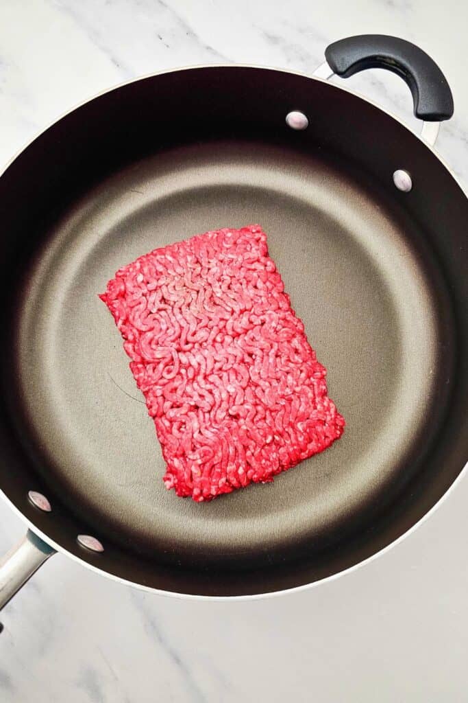 Uncooked ground beef placed in a nonstick skillet, ready to be browned on the stovetop as the first step in preparing a beef quesadillas dish. The skillet rests on a marble countertop.