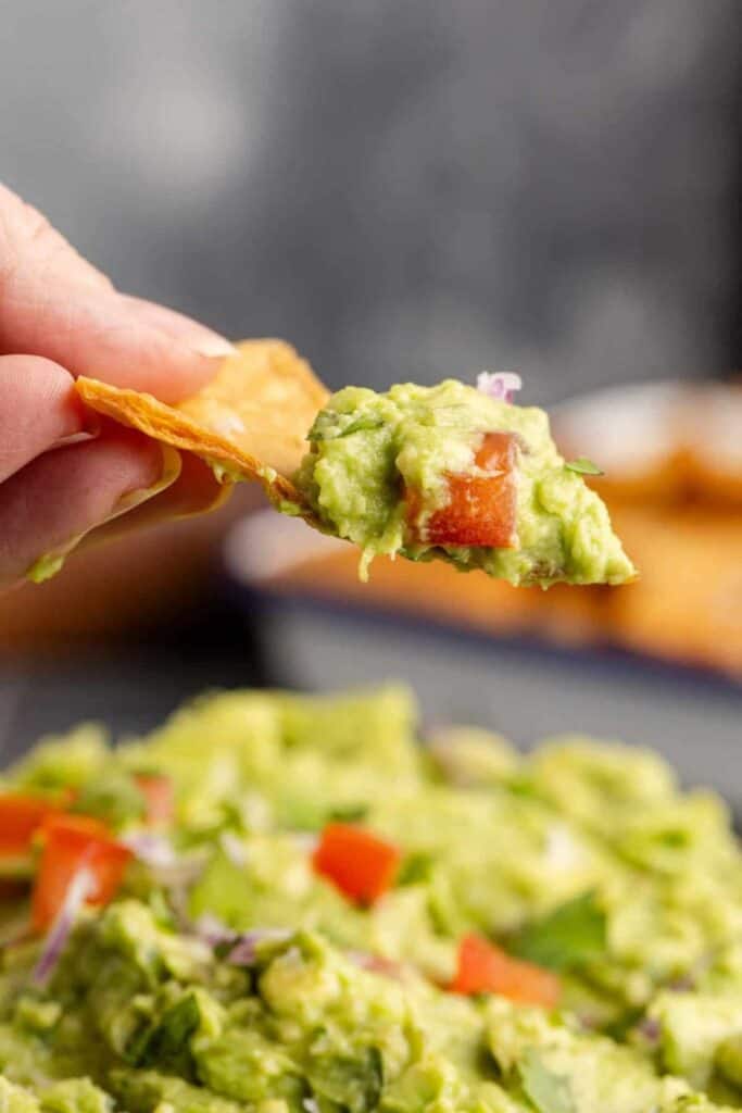 Close-up of a hand holding a tortilla chip scooping fresh guacamole, with visible chunks of tomato, red onion, and cilantro. The bowl of guacamole in the background adds a colorful, creamy contrast perfect for a party or cookout.