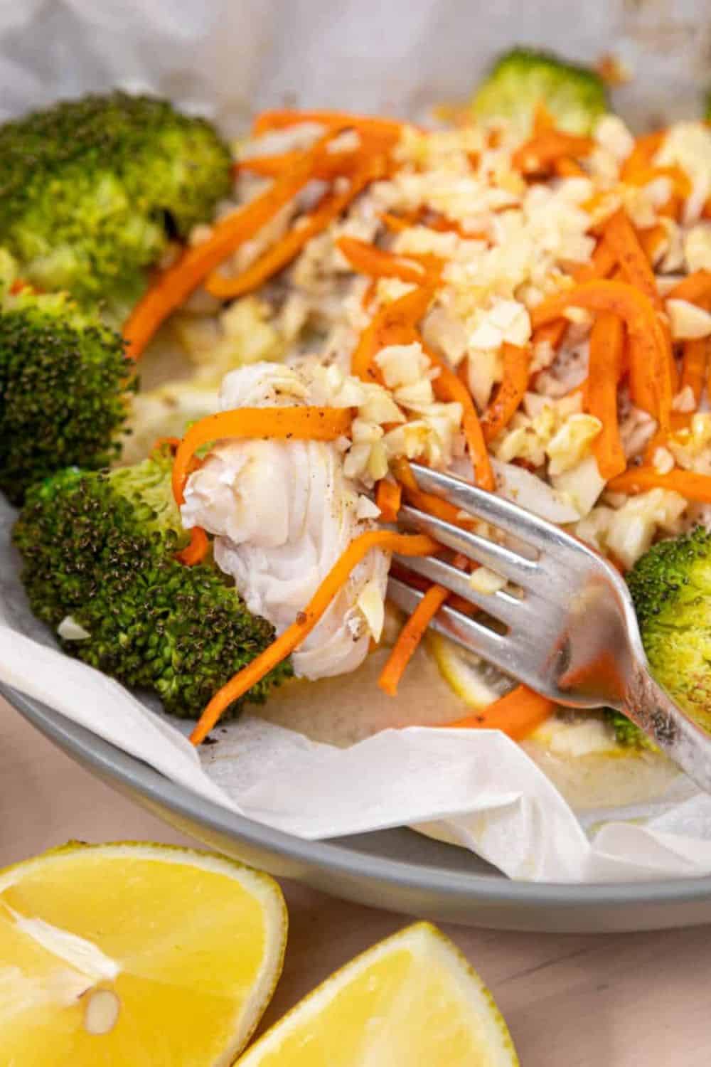 Flaky haddock baked en papillote with broccoli, shredded carrots, garlic, and lemon slices, with a fork lifting a tender piece of fish from the parchment packet. 