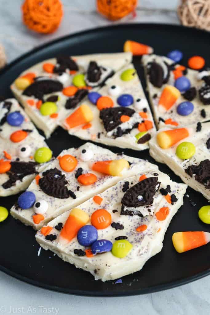 Pieces of Halloween candy bark made with a white chocolate base and topped with crushed chocolate sandwich cookies, candy corn, colorful M&M's, candy eyes, and festive orange and purple sprinkles. Served on a black plate for a vibrant and spooky sweet treat.