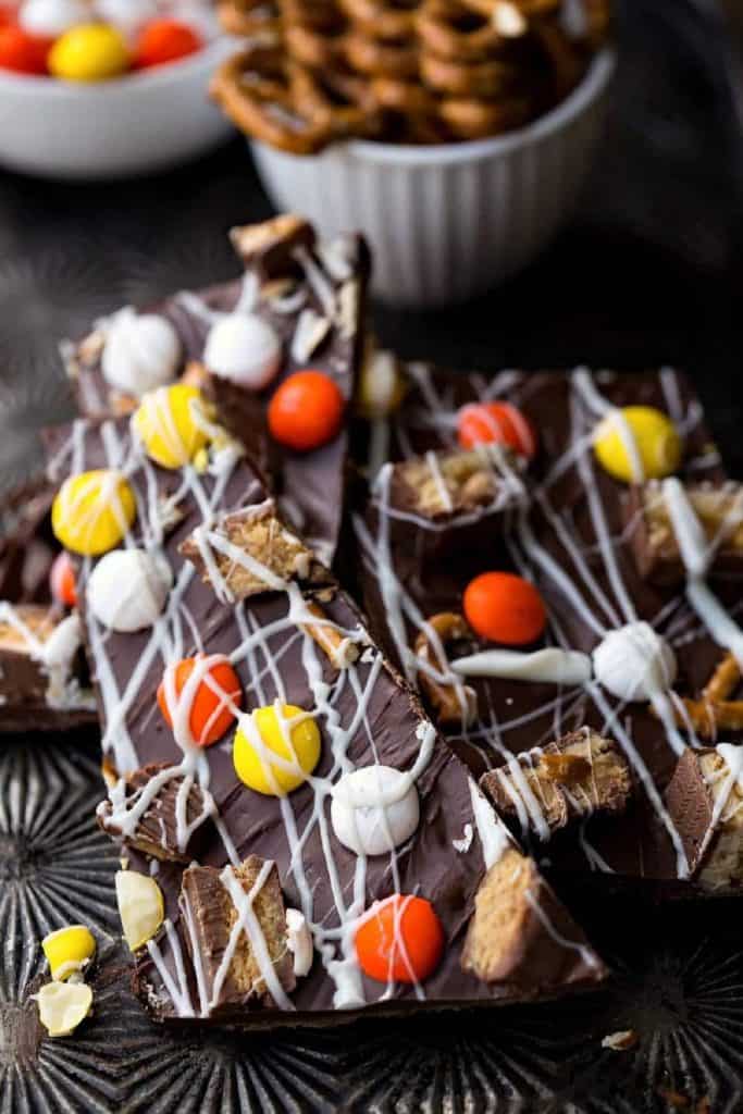 Close-up of Halloween chocolate bark topped with colorful candies, pretzel pieces, chopped peanut butter cups, and drizzled white chocolate. The bark is broken into shards and stacked on a dark textured surface with bowls of pretzels and candy in the background.