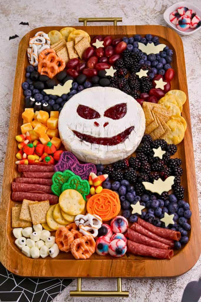 Halloween charcuterie board arranged on a wooden tray featuring a round brie wheel with a spooky Jack Skellington face made of jam, surrounded by an assortment of crackers, cheeses, cured meats, grapes, blackberries, blueberries, candy eyeballs, festive cookies, candy corn, and Halloween-shaped cheese cutouts. A colorful and creepy mix of sweet and savory snacks perfect for a themed movie night or party.