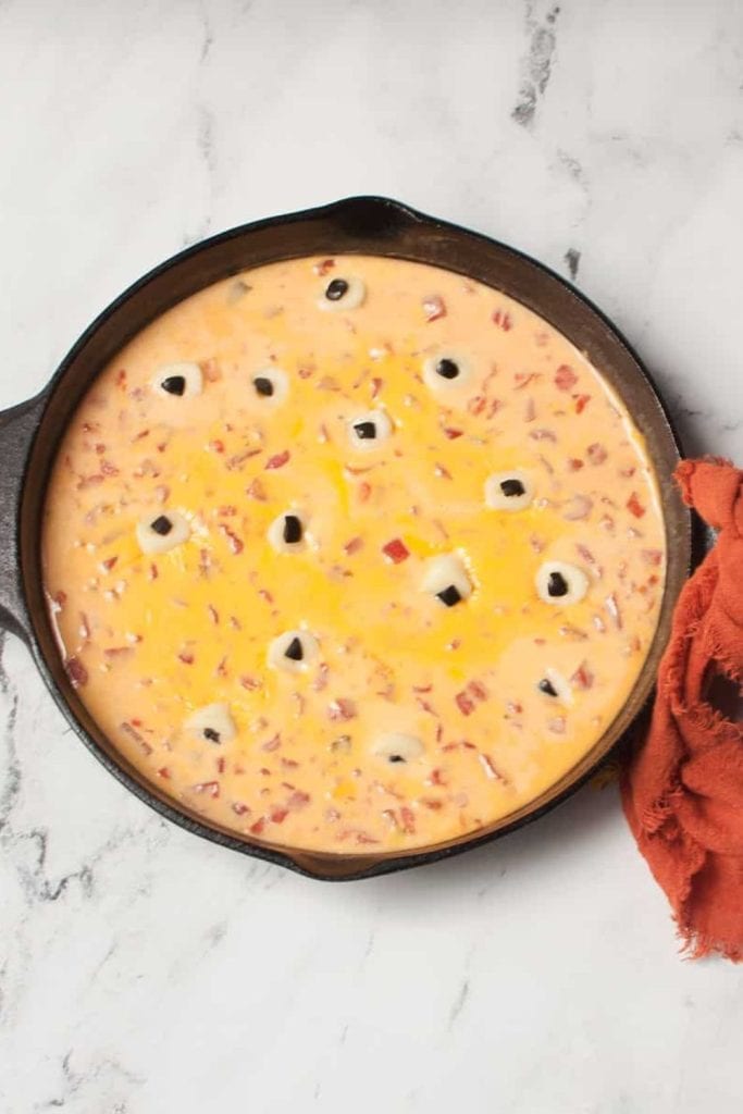Cast iron skillet filled with cheesy Halloween queso dip, speckled with diced tomatoes and topped with olive-stuffed mozzarella balls made to look like spooky eyeballs. Set on a marble surface with a burnt orange napkin for a festive fall touch.