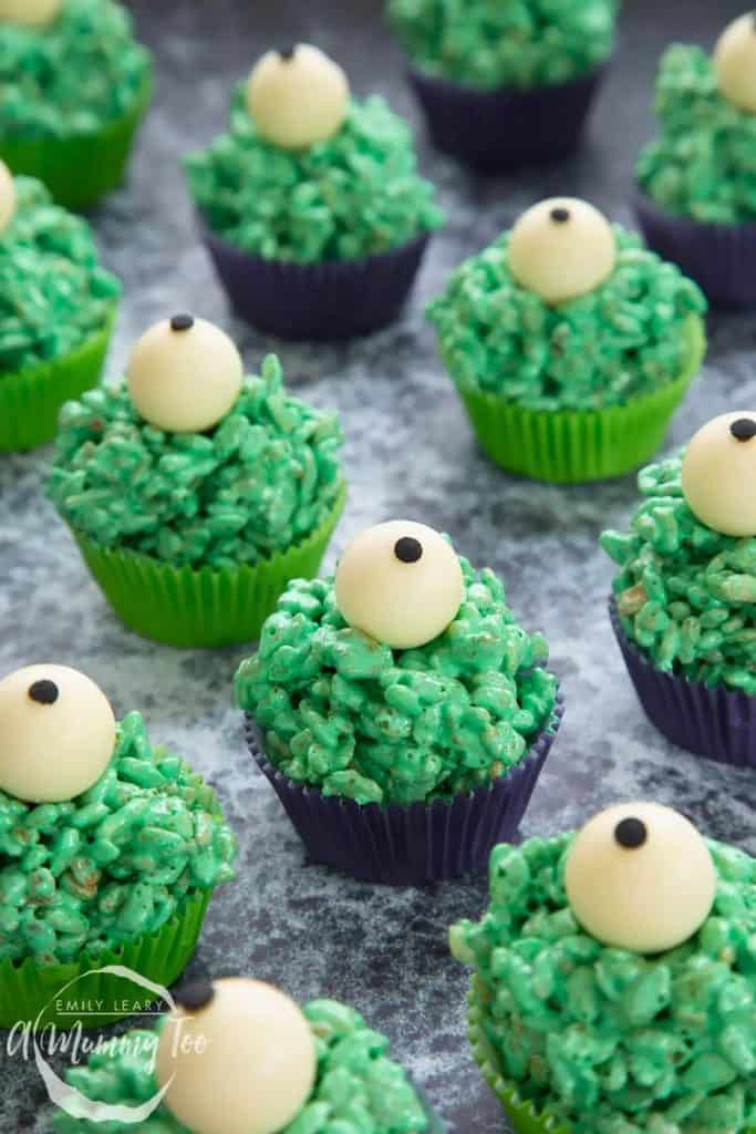 Green Monster Rice Krispies treats shaped into cupcakes with a white chocolate eyeball on top, set in green and purple paper liners on a gray surface.