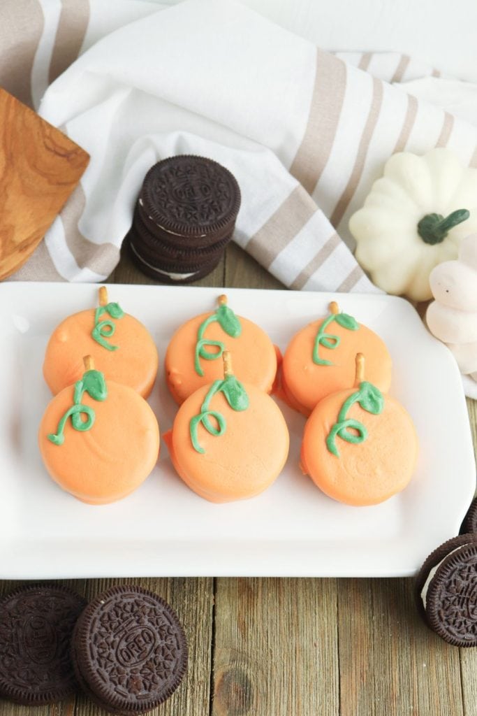 A single Oreo cookie with a pretzel stem is held above a bowl of melted orange candy, ready to be dipped for a Halloween pumpkin treat. More cookies and pretzel sticks are scattered in the background, set against a striped orange and white towel for a festive touch.